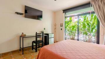 Un dormitorio con cama, escritorio, silla, televisor en la pared y vista a zonas verdes.