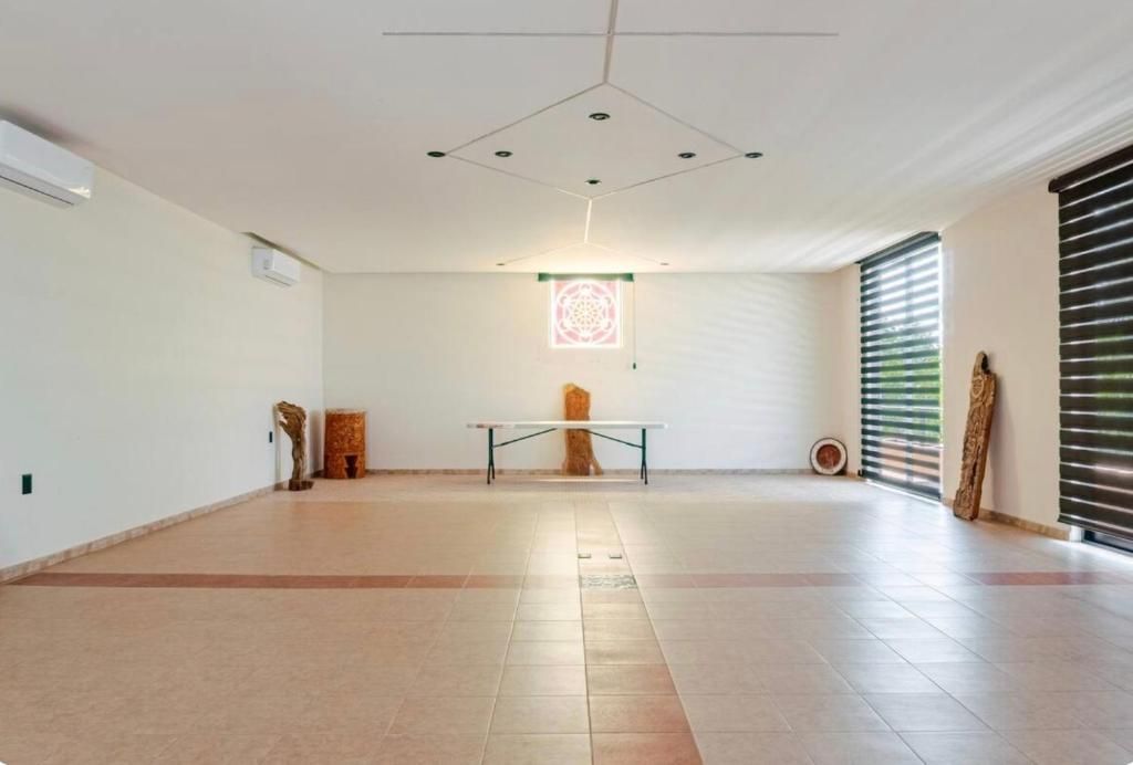 Una habitación amplia con techo alto, una mesa central y un ventanal con persianas.