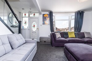 A living room with a grey sofa, a purple sofa, a staircase, a door with floral glass panels, and a window with curtains. - Spacious 4 Bed Detatched Nottingham House I Sleeps 11 I Parking for 3 I Garden (Nottingham)