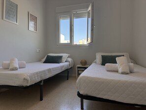 A room with two beds, each with a white duvet and pillows, a small bedside table with a globe, and a window with a view of buildings. - Apartamento Texas Life - Ole Solutions (Jerez de la Frontera)