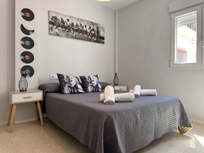 A bedroom with a bed, pillows, a nightstand, a wall-mounted picture, and a window. - Apartamento Texas Life - Ole Solutions (Jerez de la Frontera)