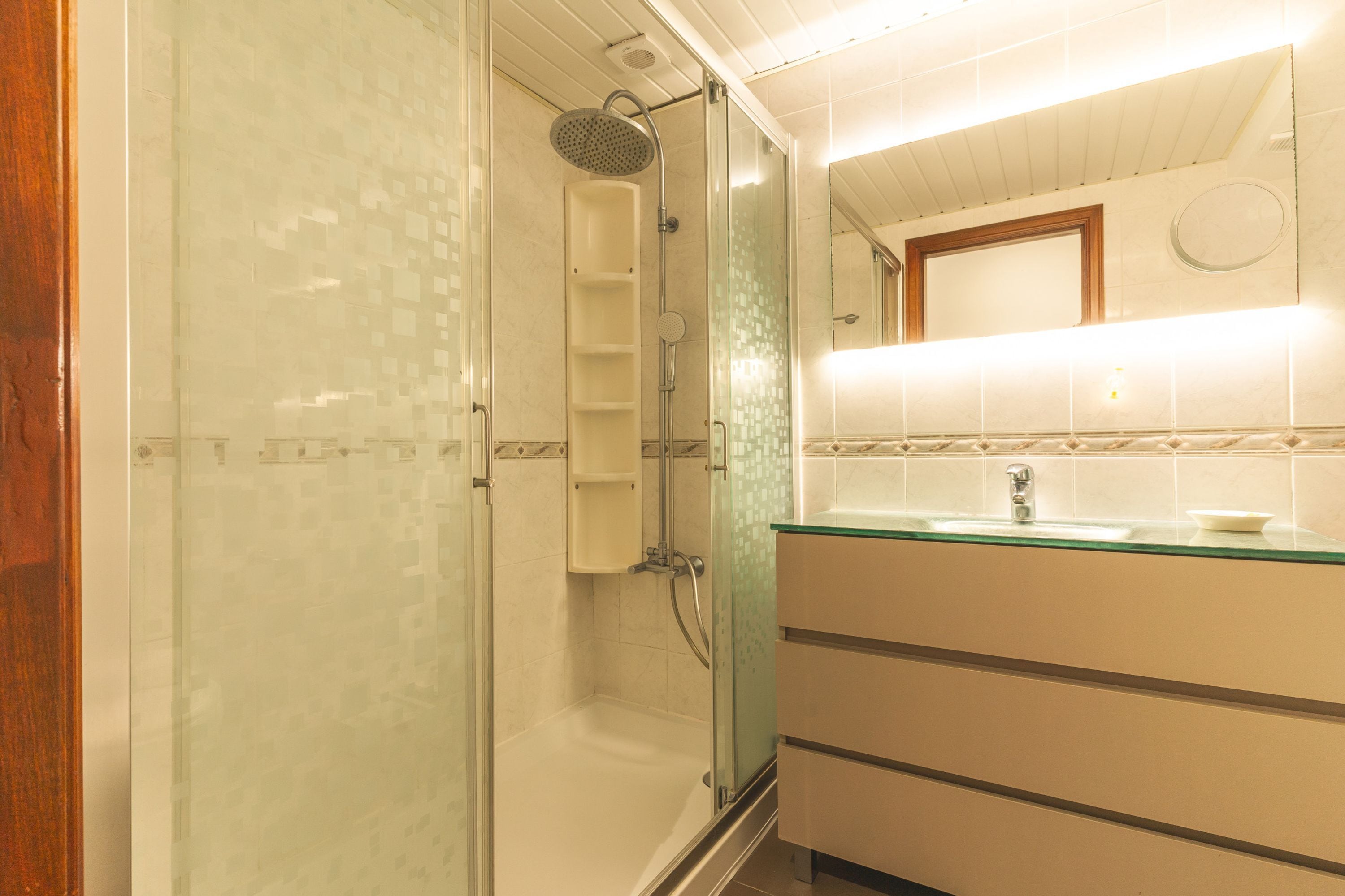Un baño con cabina de ducha de vidrio, lavamanos con estructura de vidrio y un espejo sobre el lavamanos.