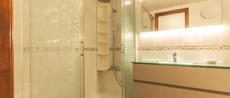 Un baño con cabina de ducha de vidrio, lavamanos con estructura de vidrio y un espejo sobre el lavamanos.