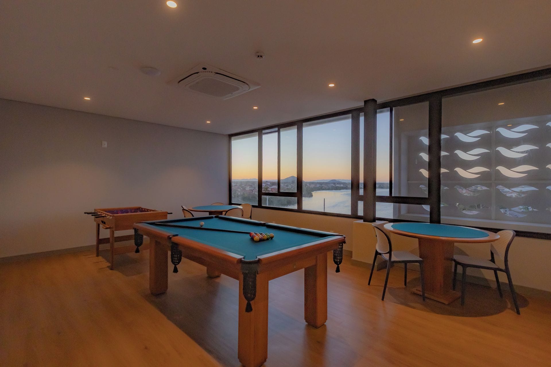 Mesa de billar y mesa de fútbolín en una habitación con ventanas amplias y vista a un paisaje urbano.