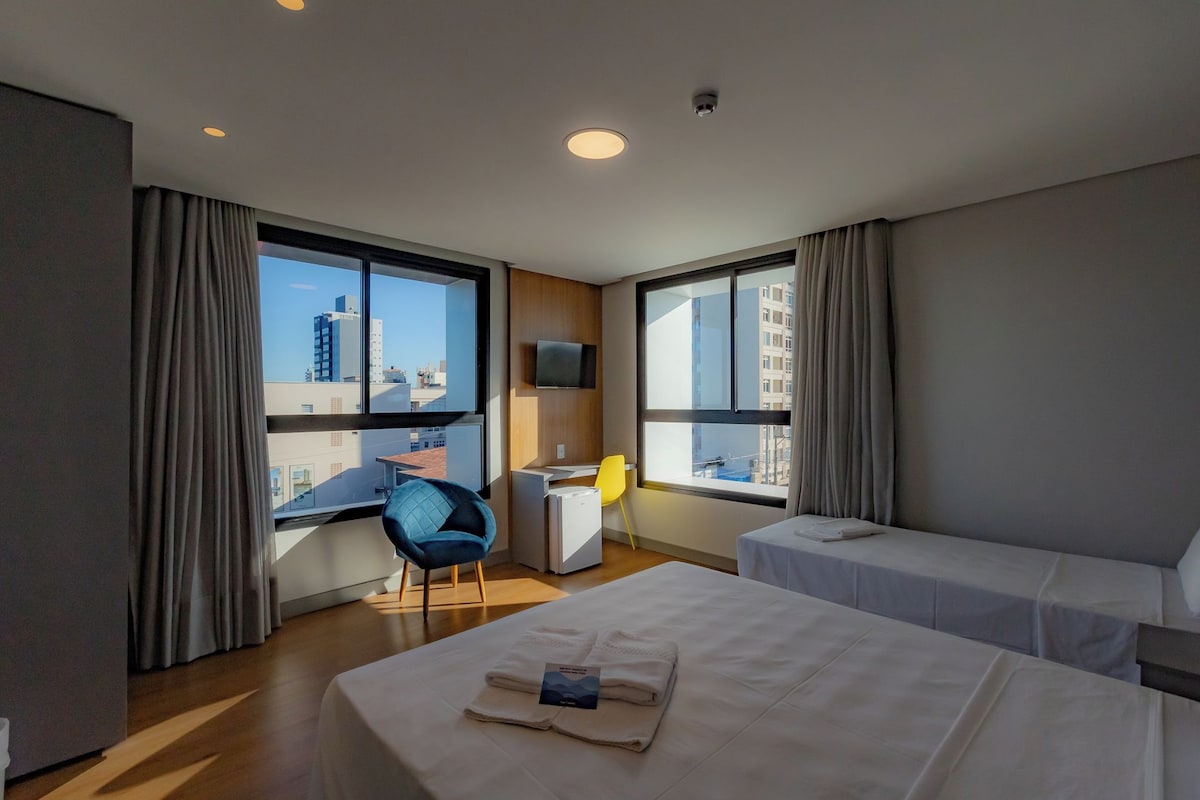 A hotel room with two beds, a desk, a chair, and large windows offering a city view.