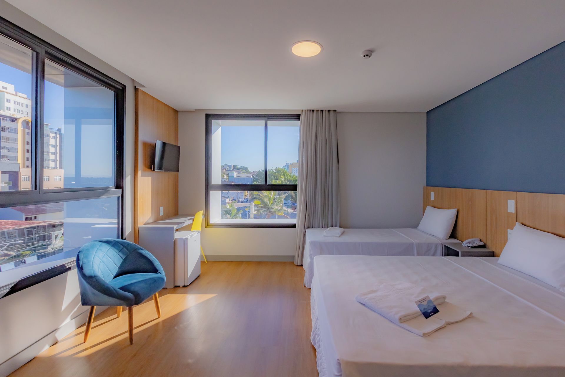 A hotel room with two beds, a desk, a chair, a TV, and a window with a view of buildings.