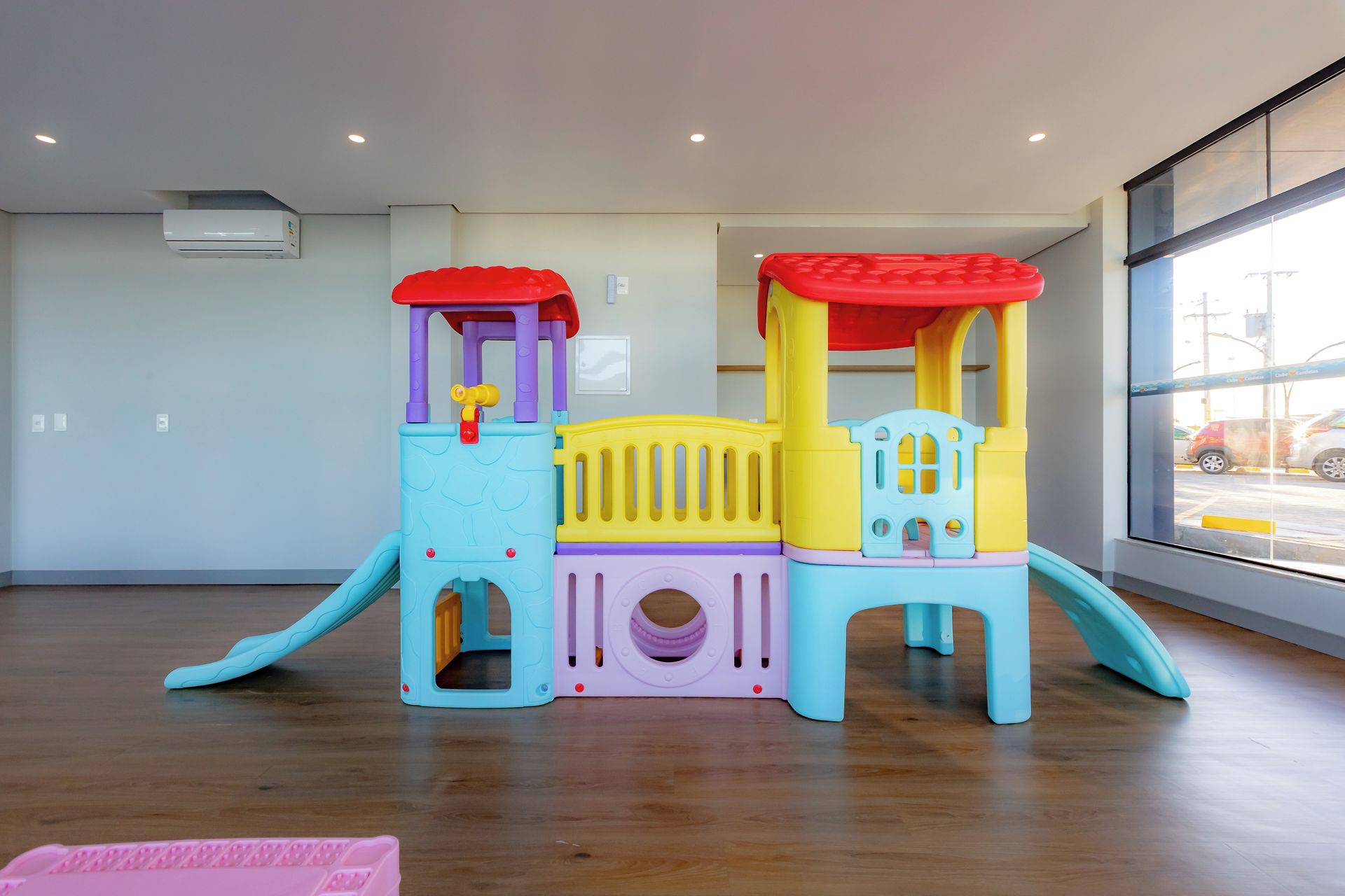 Una estructura de juegos infantiles en una habitación con suelo de madera, una ventana con vista a la calle y un equipo de aire acondicionado en la pared.