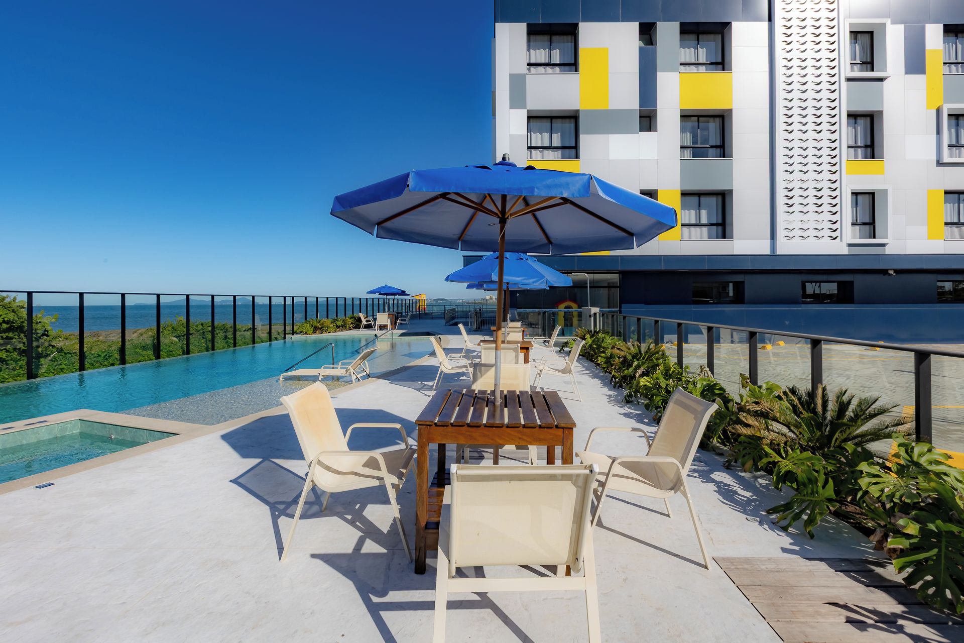 A rooftop pool area with lounge chairs and umbrellas, overlooking a body of water and a modern building.