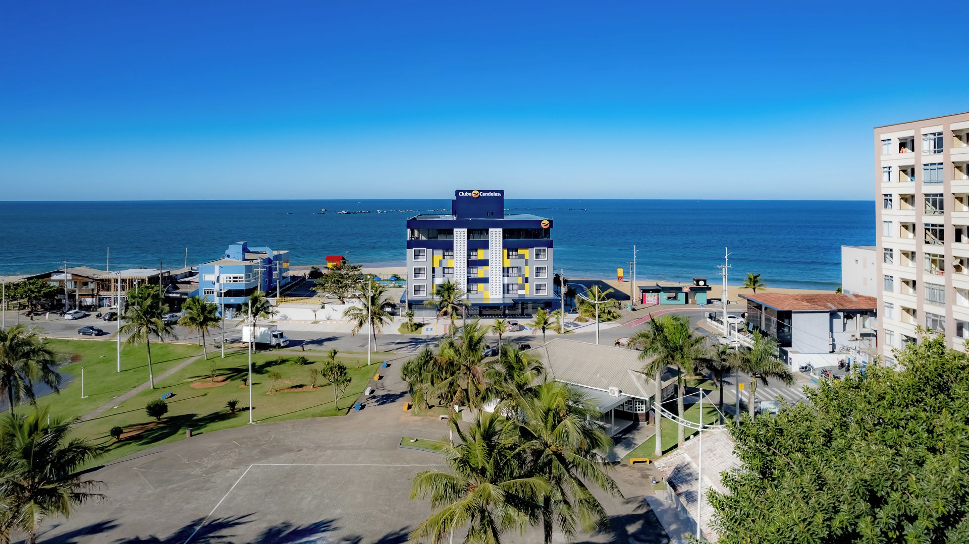 A hotel with a central building and a parking lot, surrounded by palm trees and a view of the ocean.