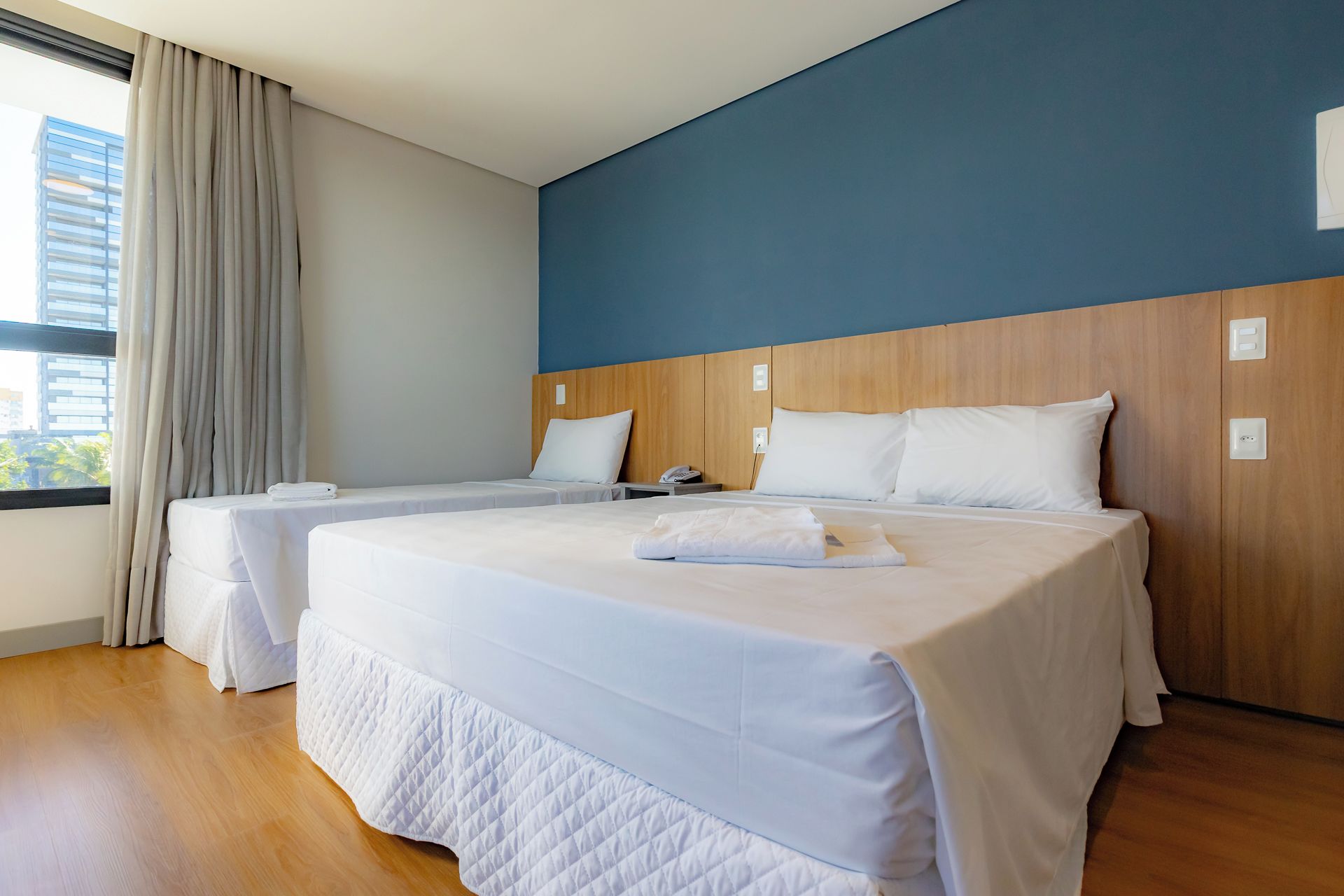 A hotel room with two beds, a wooden headboard, a window with curtains, and a view of buildings outside.