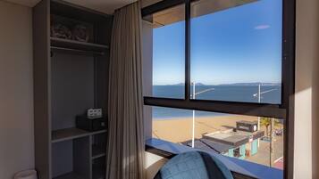 A room with a large window overlooking a beach, a bookshelf, and a chair.