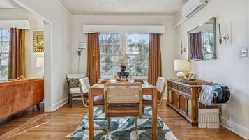 A dining area with a wooden table, chairs, and a rug. There is a sofa and a cabinet with a mirror and a lamp.