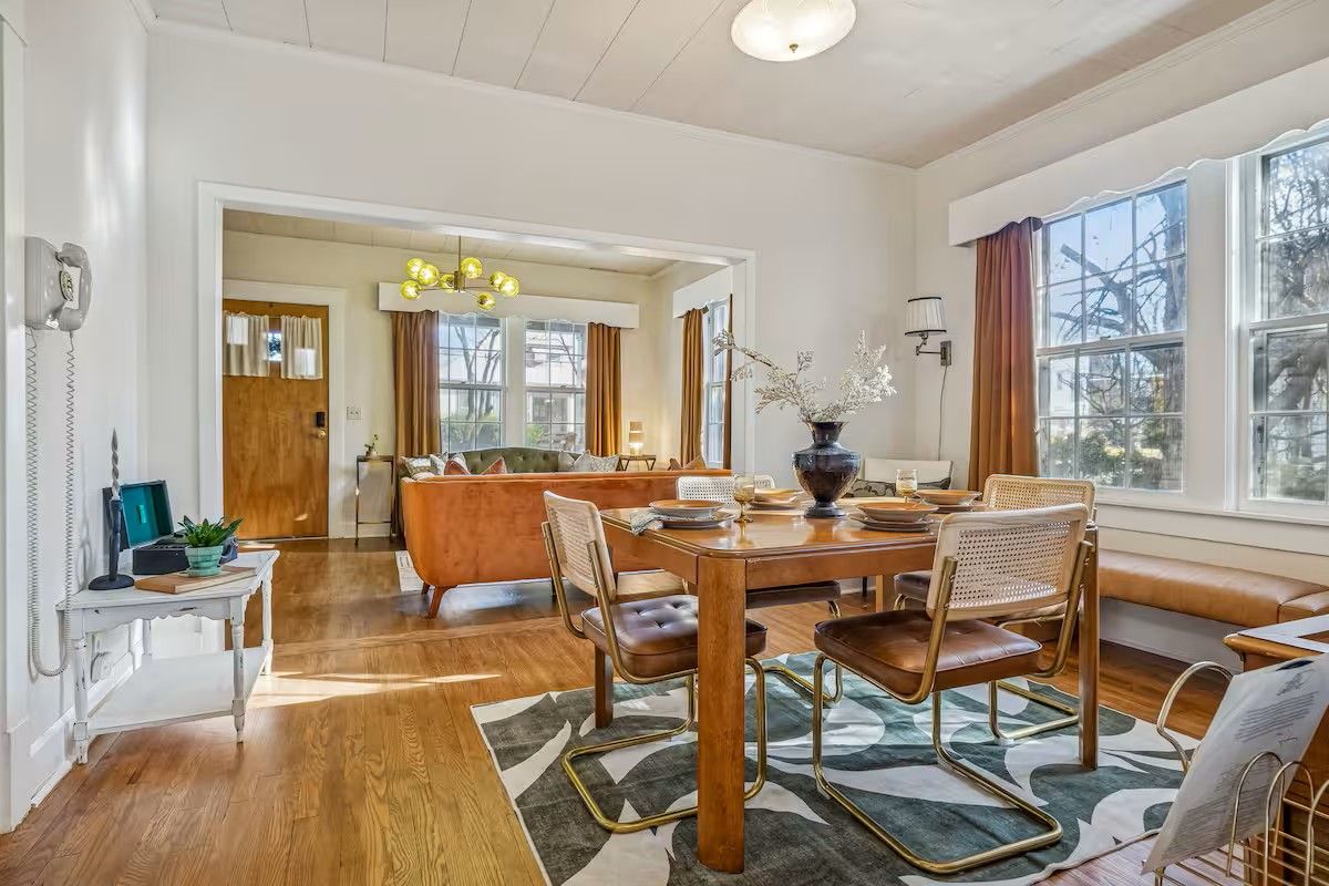 Un coin repas avec une table en bois, des chaises et un tapis. Il y a une fenêtre avec des rideaux et une vue sur les arbres à l’extérieur.