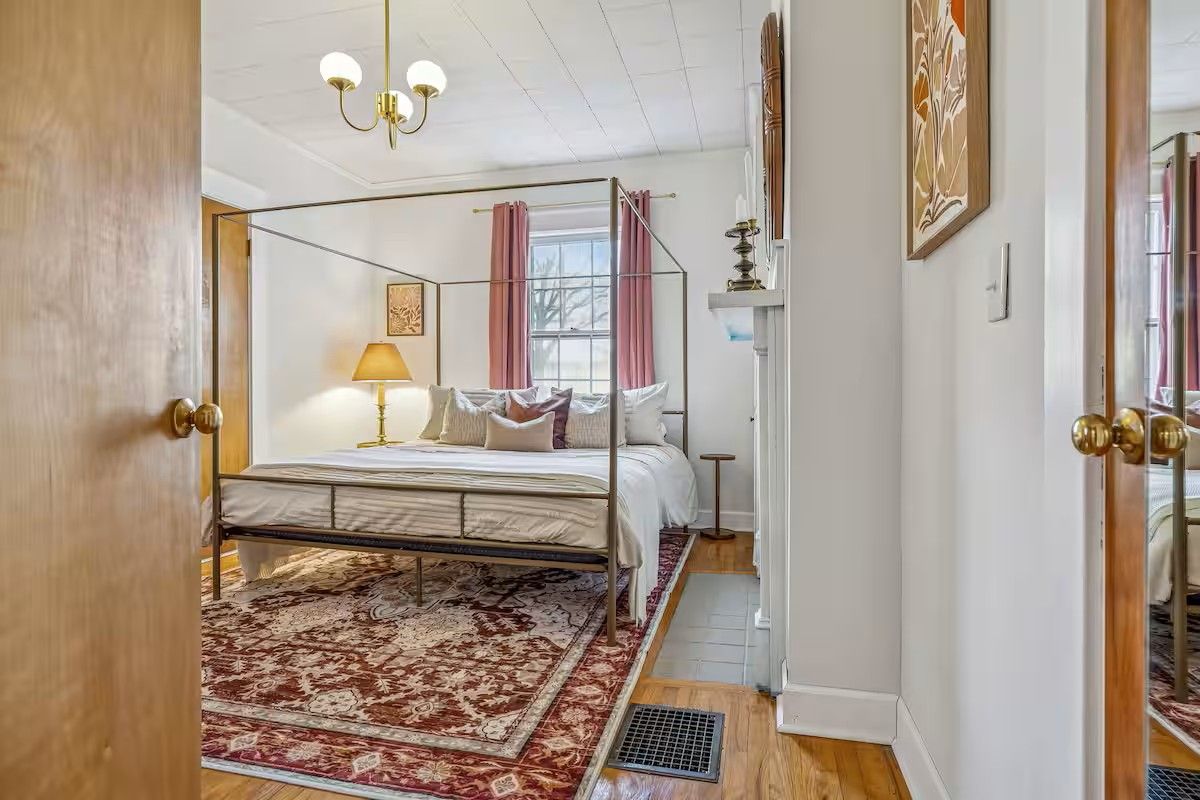 A bedroom with a four-poster bed, a red patterned rug, a bedside lamp, and a window with pink curtains.