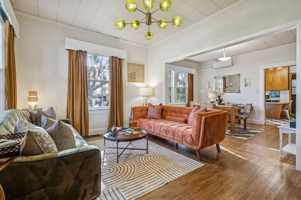 A spacious living room with a sofa, a coffee table, and a chandelier. There is a dining area with a table and chairs, and a kitchen area with cabinets and a countertop.
