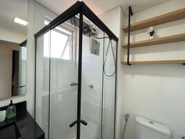 Casa de banho com cabine de chuveiro em vidro, sanita, lavatório com dispensador de sabonete e prateleira com um vaso de flores.