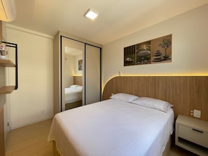Quarto de hotel com cama, mesa de cabeceira e prateleira fixada na parede com planta em vaso.