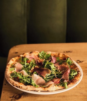 A pizza with greens and prosciutto on a white plate on a wooden table. - The Lauriston Pub & Guest Rooms (London)