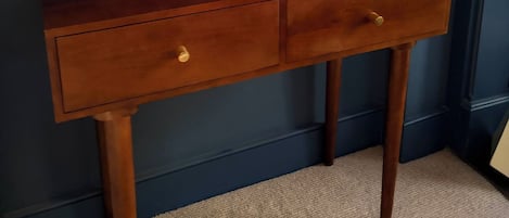 A wooden desk with two drawers against a blue wall, a framed picture above it, and a dark cabinet to the right.