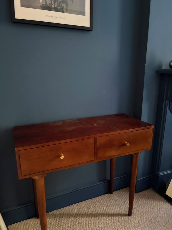 A wooden desk with two drawers against a blue wall, a framed picture above it, and a dark cabinet to the right.