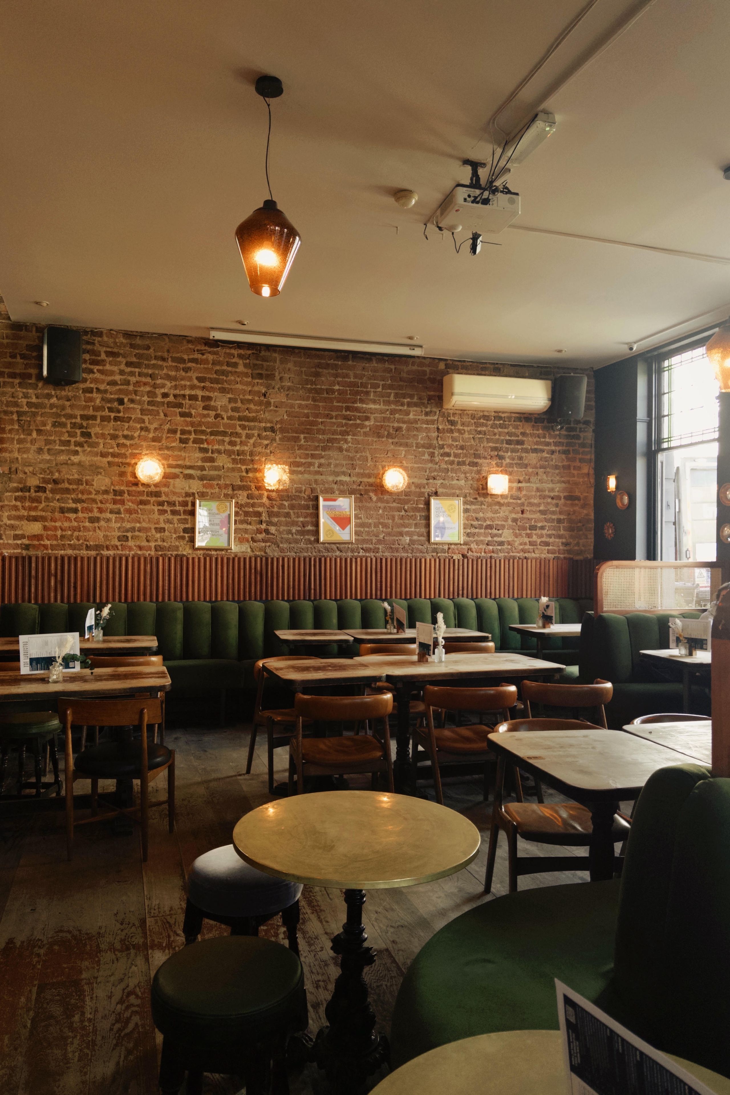 A restaurant interior with brick walls, green booths, wooden tables, and hanging pendant lights.