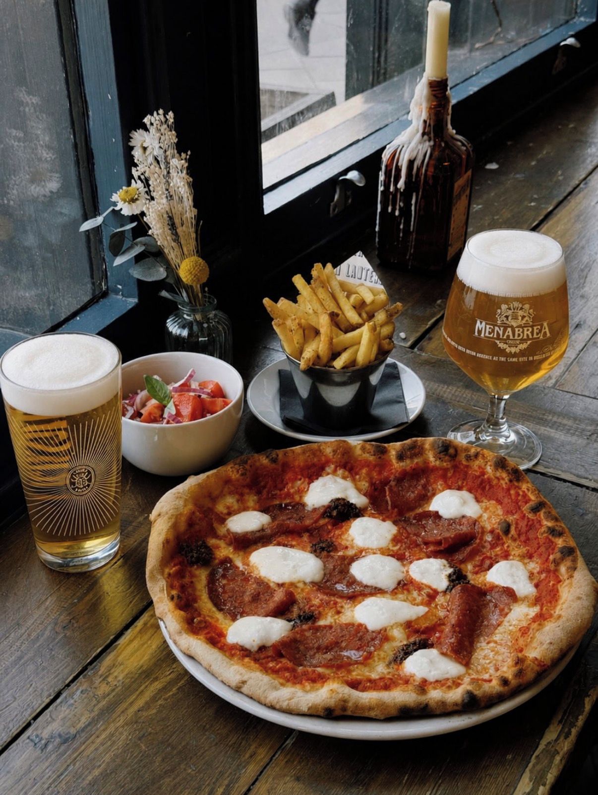 Pizza, een biertje, een saladekom en een fles saus op een houten tafel.
