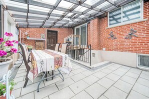 Een overdekt terras met een eettafel, een bakstenen muur en een raam.