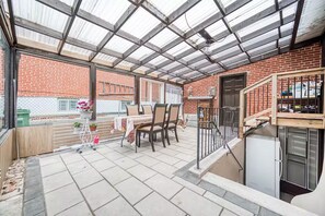 Een overdekt terras met een bakstenen muur, een tafel met stoelen, een kleine tuin en een trap.