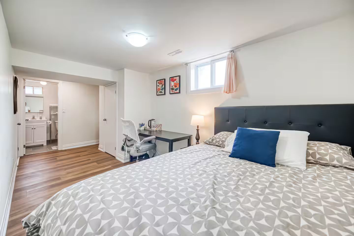 Ein Schlafzimmer mit einem großen Bett, einem Schreibtisch und einem Stuhl. Es gibt ein Fenster mit Vorhängen, eine Deckenleuchte und eine Tür zu einem Badezimmer.