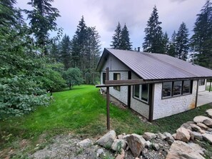 A single-story building with a metal roof, surrounded by a well-maintained lawn and dense forest. - Salmon Valley Guest Ranch - Cozy 3 bedroom cottage (Columbia-Shuswap)