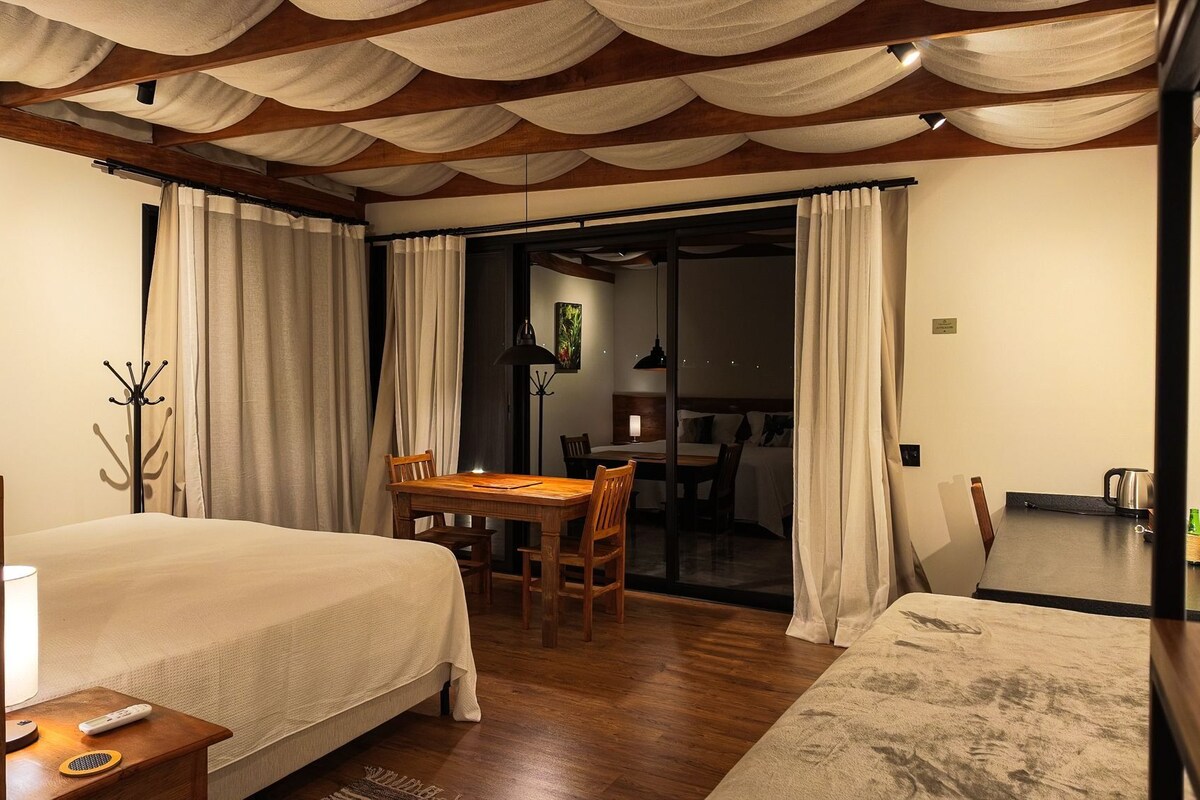 Ein Hotelzimmer mit einem Bett, einem Esstisch mit Stühlen, einem kleinen Tisch mit einer Lampe und Blick auf den Gastbereich.