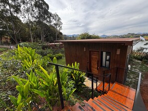 Una cabaña de madera con una escalera que lleva a una terraza, rodeada de exuberante vegetación y vistas a colinas distantes.