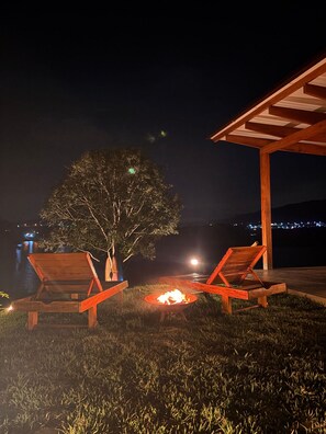 Un espacio al aire libre con asientos nocturnos, dos sillones de madera, un pequeño hogar para fuego y una estructura de pérgola.