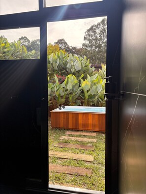 Vista a través de una puerta que da a un patio trasero con jacuzzi, terraza de madera y exuberante vegetación.