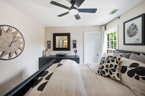 Un dormitorio con cama, ventilador de techo, reloj de pared, espejo y ventana con cortinas.