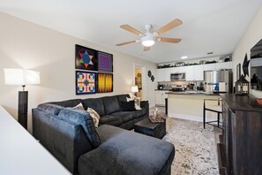 A living room with a sectional sofa, a ceiling fan, and a kitchen in the background.