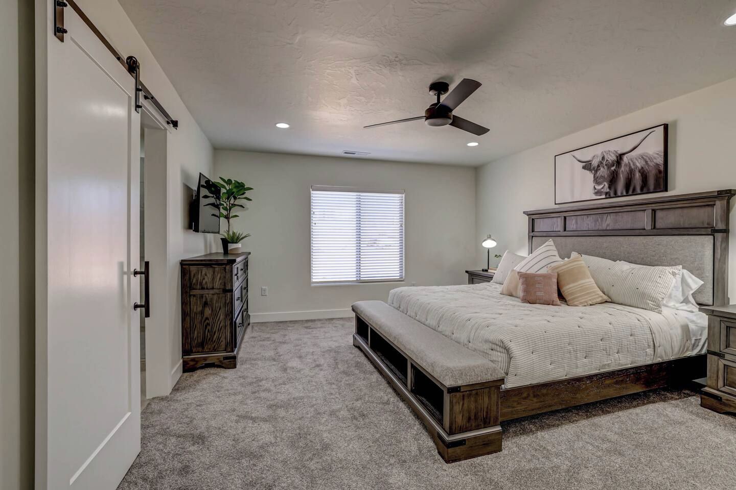 A bedroom with a bed, a bench, a ceiling fan, a window with blinds, and a wall-mounted plant.