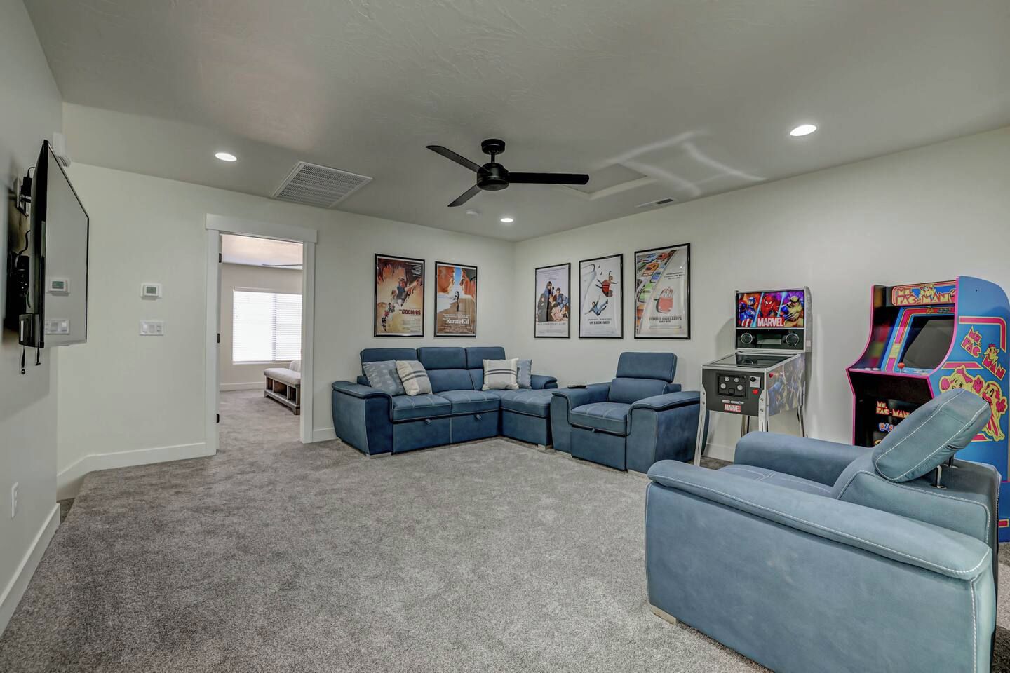 A living room with a sectional sofa, a ceiling fan, wall-mounted posters, and a gaming console.