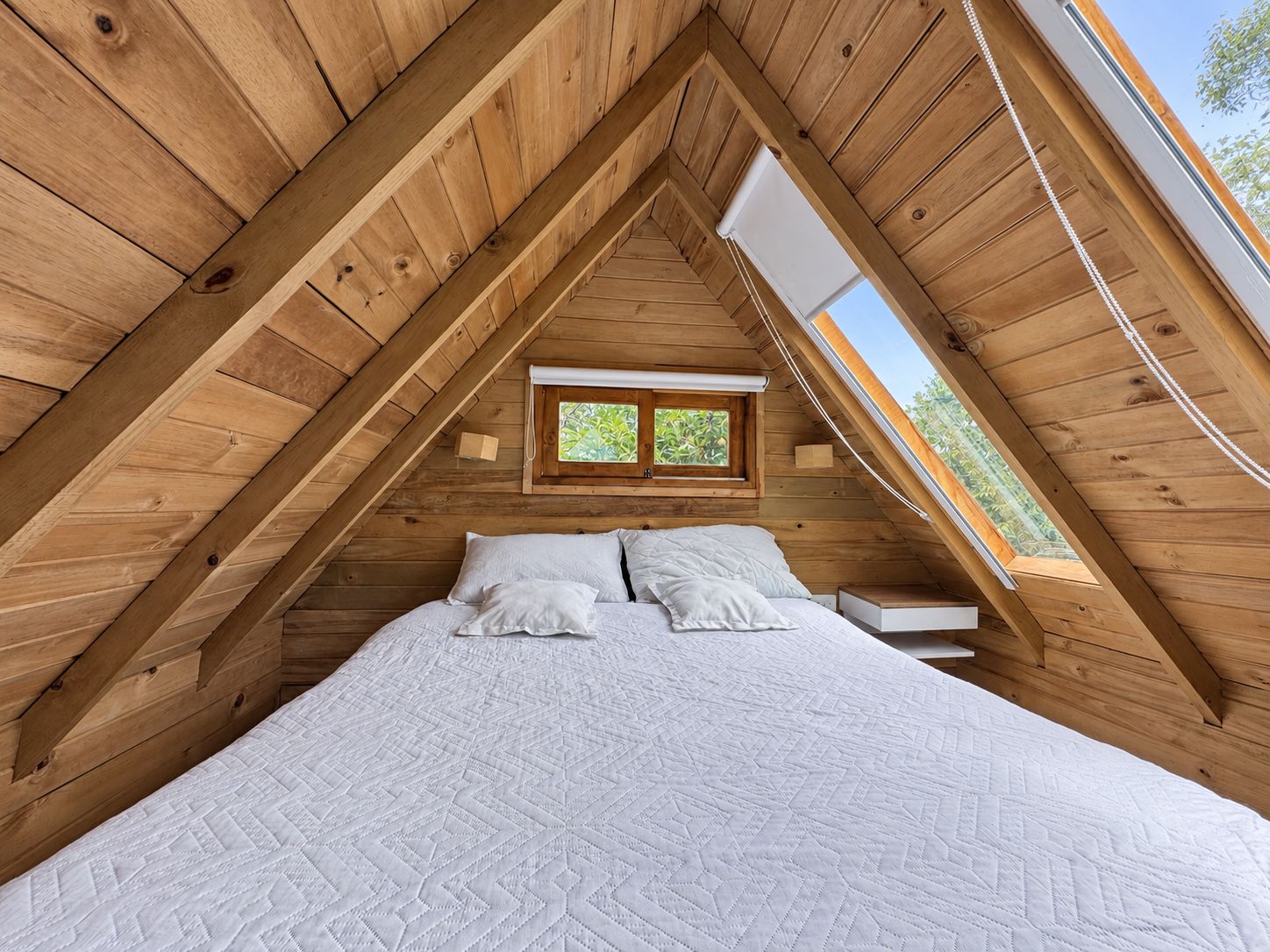 A bedroom with a sloped ceiling, wooden walls, a bed with white bedding, and a window with a view of greenery.