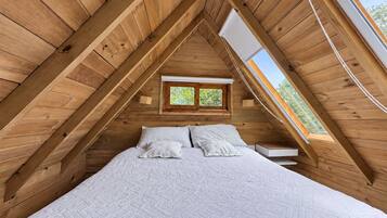 A bedroom with a sloped ceiling, wooden walls, a bed with white bedding, and a window with a view of greenery.