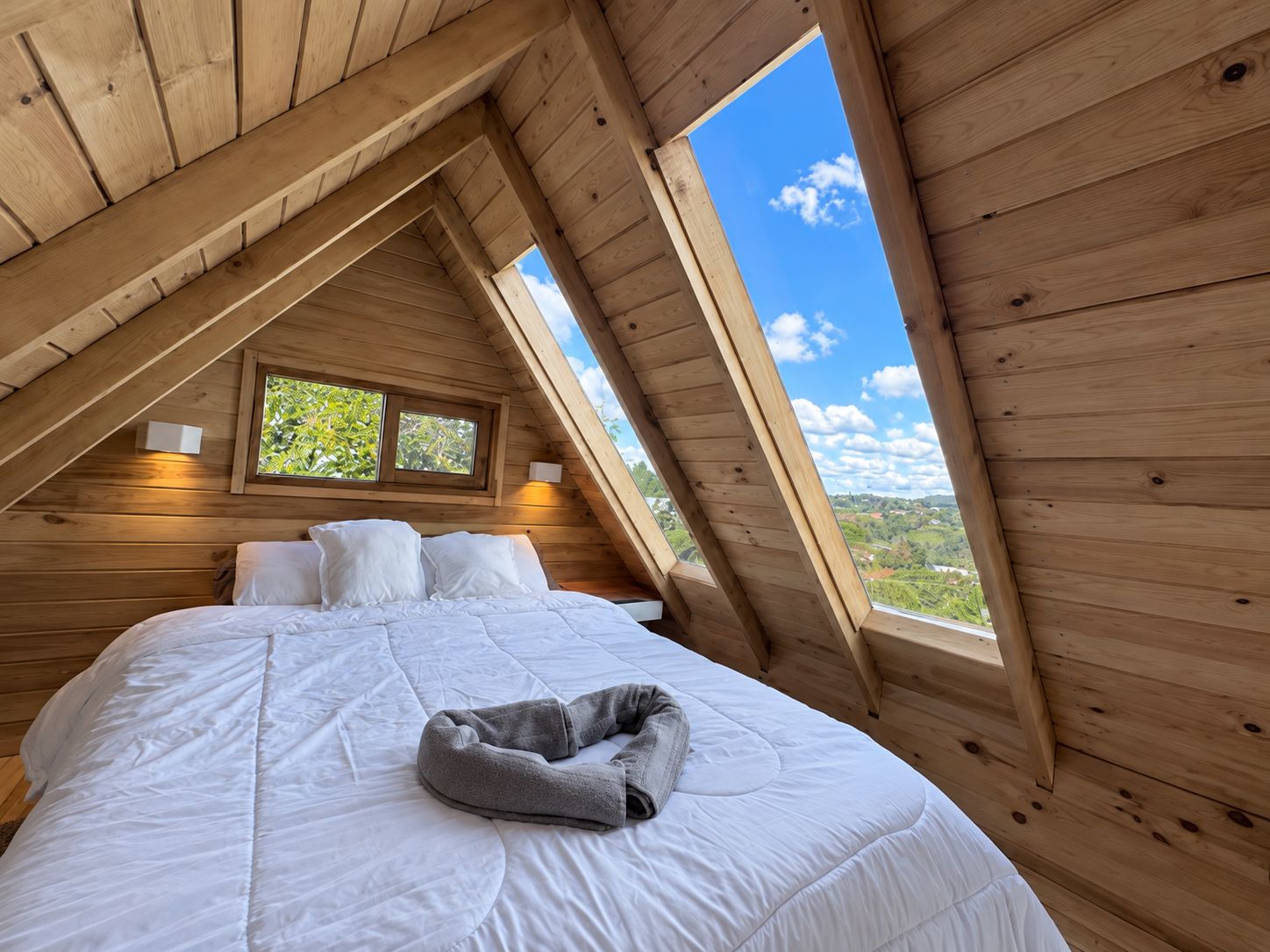 A bedroom with a sloped ceiling, a bed with white bedding, two bedside lamps, and a window with a view of greenery.