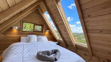Een slaapkamer met een schuine kap, een bed met witte beddengoed, twee nachtlampjes en een raam met uitzicht op groen.