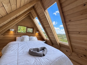 A bedroom with a sloped ceiling, a bed with white bedding, two bedside lamps, and a window with a view of greenery.