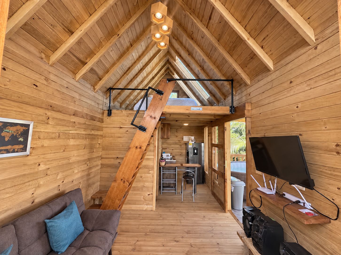 Interior de una cabaña de madera con cama en un altillo, kitchenette y un televisor sobre una repisa de madera.