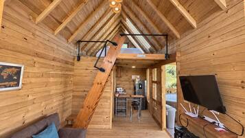 Interior de una cabaña de madera con cama en un altillo, kitchenette y un televisor sobre una repisa de madera.