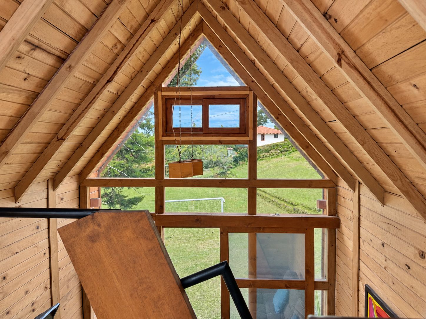 A wooden interior with a triangular roof structure, a window with a view of a green landscape, and a wooden bench.