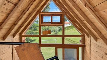 A wooden interior with a triangular roof structure, a window with a view of a green landscape, and a wooden bench.