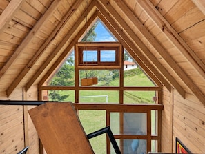 Un interior de madera con estructura de techo triangular, una ventana con vistas a un paisaje verde y un banco de madera.