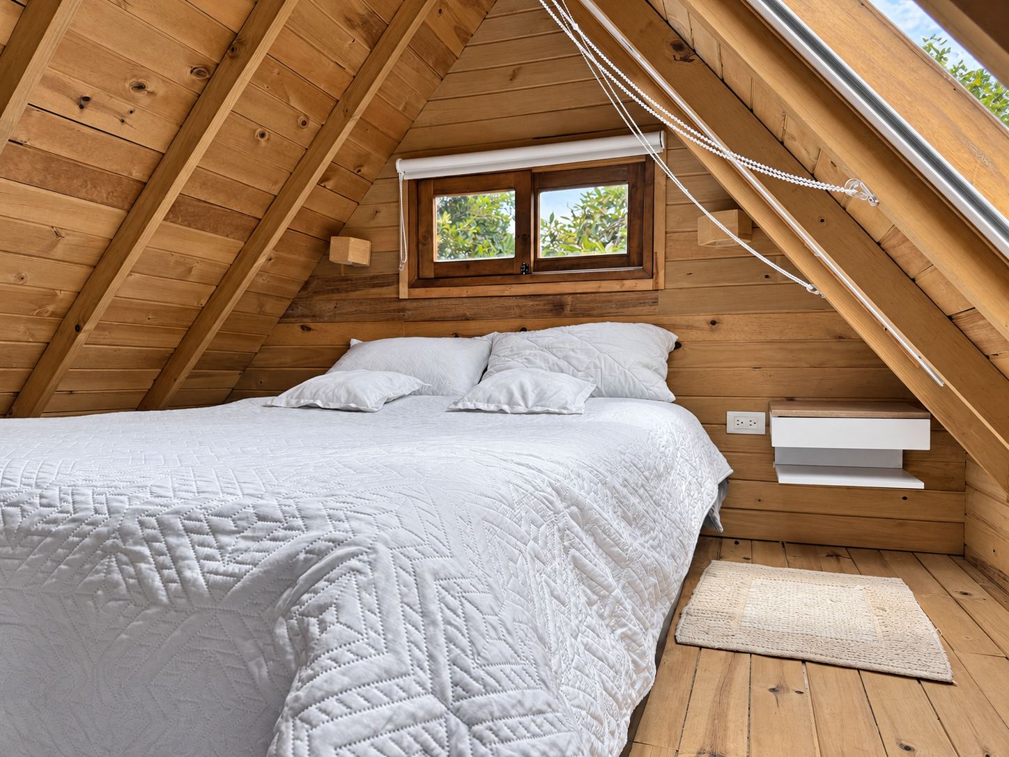 Un dormitorio con techo inclinado de madera, una cama con ropa de cama blanca, un ventanal pequeño y una mesita de noche.