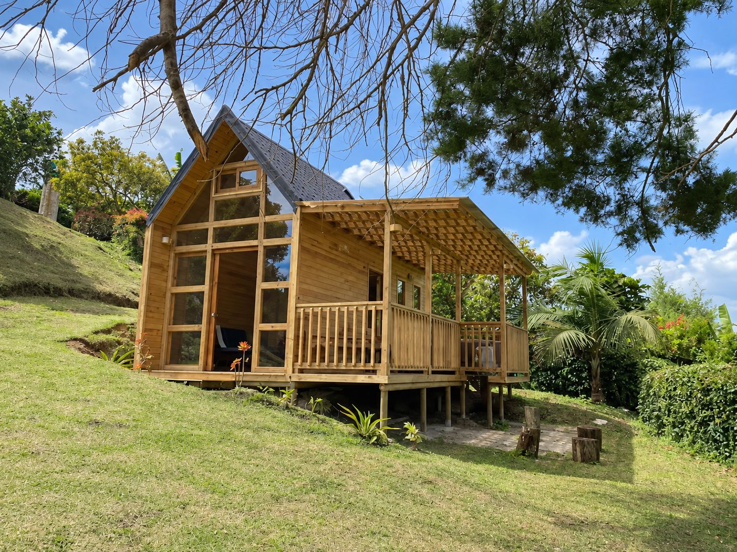 Una cabaña de madera con balcón, ubicada en una ladera cubierta de hierba y árboles al fondo.